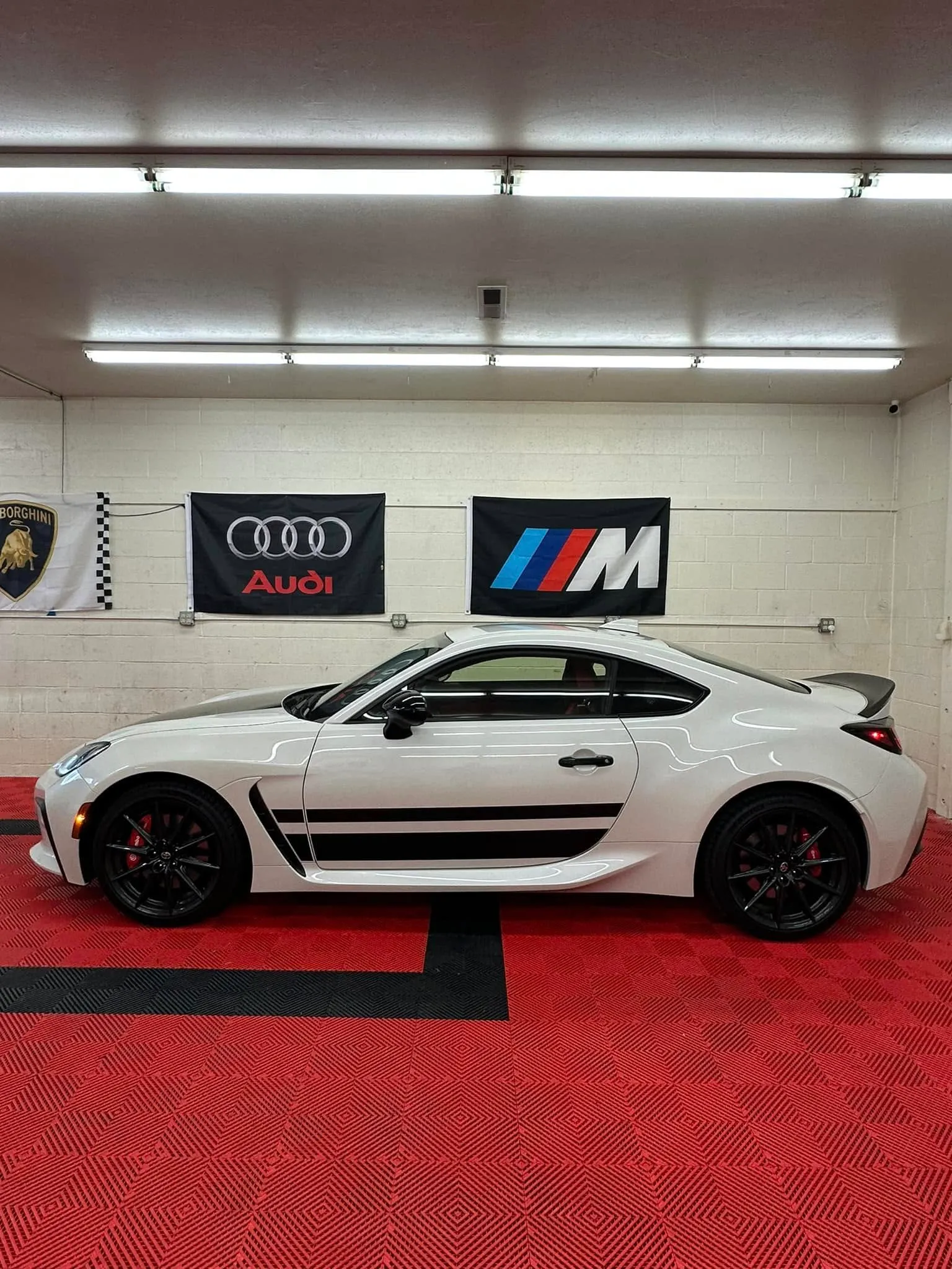 White Porsche 718 Cayman with black side stripes inside the Redline Tinting install bay with Audi and BMW M banners on the wall